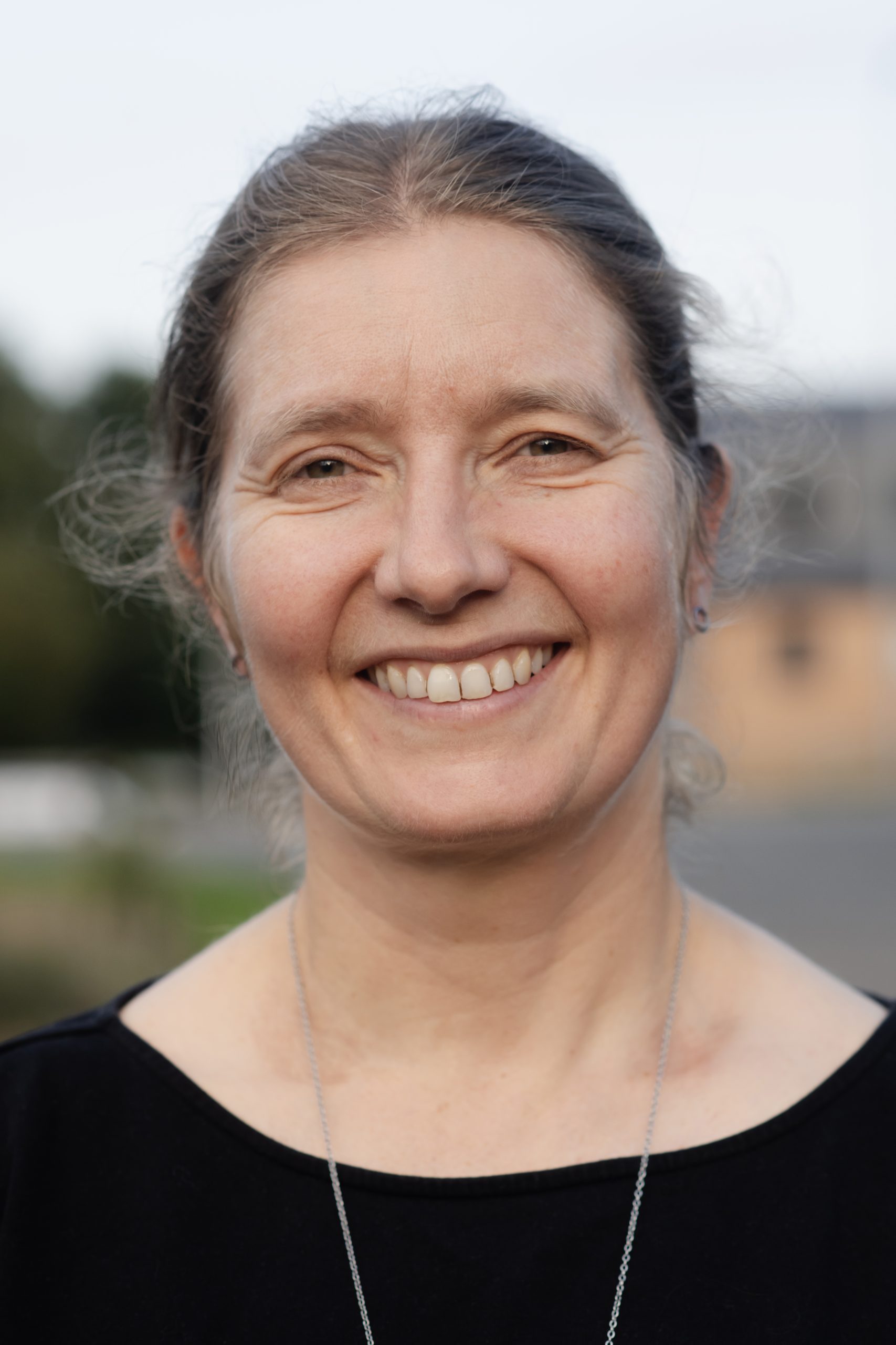 Camilla, a white woman with her blonde hair pulled back behind her neck, looks at the camera. She is smiling and is wearing a black top and a delicate gold chain.