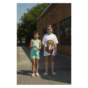 Two young girls stand together. One has a cartoon character on her t-shirt. The other is wearing a green vest and shorts.