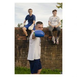 Photograph of 3 male boxers. 2 sit on a wall. One in the forground rasies a punch towards the camera