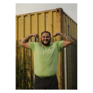 A man stands in front of a yellow shipping container. He is wearing a lime green t-short and is flexing his arm muscles and smiling.