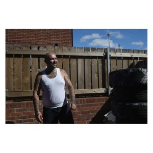 A man in a white vest stands infront of a fence next to a stack of tyres. He is smiling