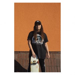 A young woman wearing a black t-shirt stands in front of an orange wall. She is resting her skate board against her legs.