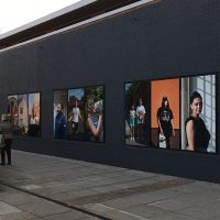 An outdoor exhibition of large portraits of people, installed on a grey wall. A group of women stand looking at the photos.