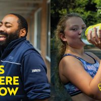 A composite image of a black man in q blue fleece laughs. A white teenager drinks from a bottle of lemonade. The words "Us Here Now" are in yellow in the bottom left corner
