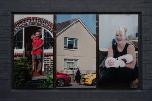 Installation shot of Us Here Now - 3 larger than life photographs showing a man in his doorway, a woman between two cars and a female boxer smiling