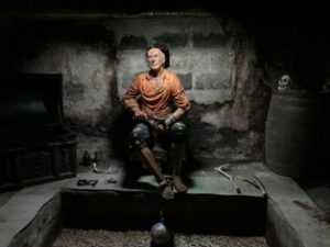 A model of a pirate with a ball and chain, sits on a chair in a cellar.