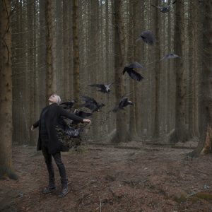 A white man in a forest, dressed in black, leans backwards with his arms open. Crows fly upwards behind him.