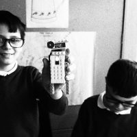 Two boys wearing glasses. One looks at the camera, smiling and holds up his robot made from lego.