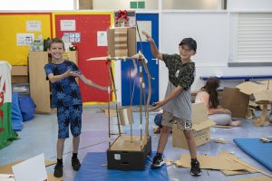 Two boys present a construction made from cardboard and carboard tubes, which is taller than they are.