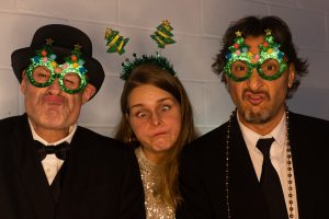 A group of three - two men wear green christmas glasses, the woman in the middle wears a a head band with christmas trees on it