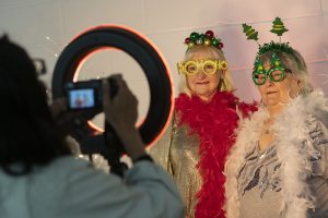 A woman with a red feather boa, yellow glasses and holly in her hair has her photo taken