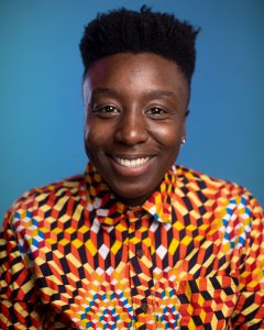 A portrait photograph of Azara - a black person with short hair wearing a yellow/ orange and black patterned shirt against a blue background