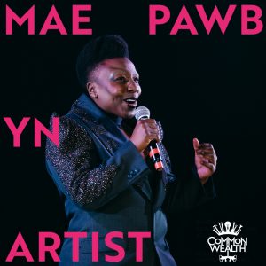 The words "MAE PAWB YN ARTIST" are laid on top of an image of a black person with short hair, wearing a blue sparkly suit, singing into a microphone