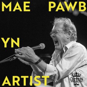The words "Mae Pawb yn artist" in blue are laid on top of a black and white image of a white man singing really loudly into a microphone