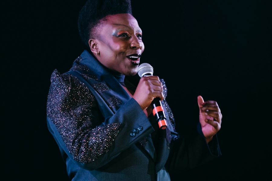 An image of a black person with short hair, wearing a blue sparkly suit, singing into a microphone