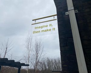 A sign, hung outside which says in yellow "Imagine it, then make it"