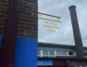A sign hangs of the side of a building. In yellow it says "all of us could build a city"