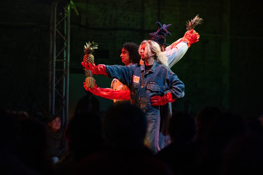 Three people wearing denim boiler suits and red gloves holding pineapples
