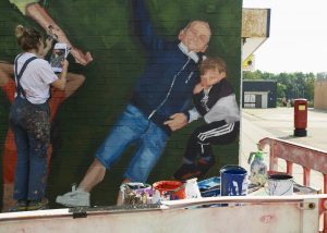 A close up of the mural showing the artist at work, beside a painted man with his small child