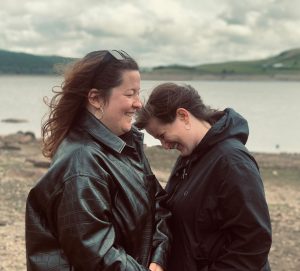 Two women in black jackets face towards each other. There is a lake and mountains in the background.