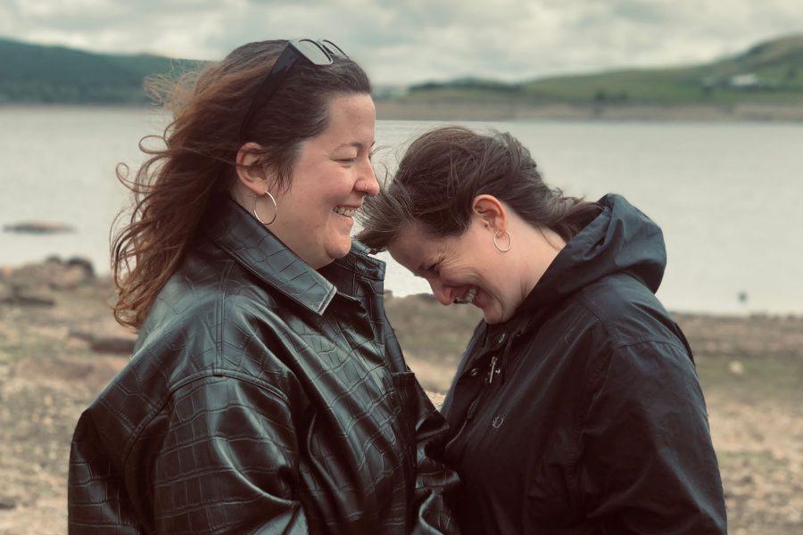 Two women in black jackets face towards each other. There is a lake and mountains in the background.