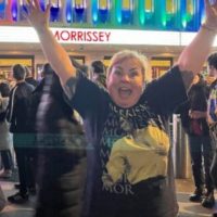 A photo of a woman with her arms in the air- she's celebrating! She is outside a music venue with people in the background and the word "Morrissey" lit up.