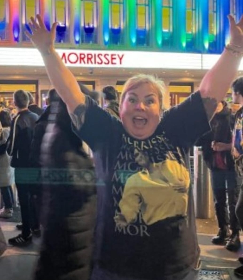 A photo of a woman with her arms in the air- she's celebrating! She is outside a music venue with people in the background and the word "Morrissey" lit up.
