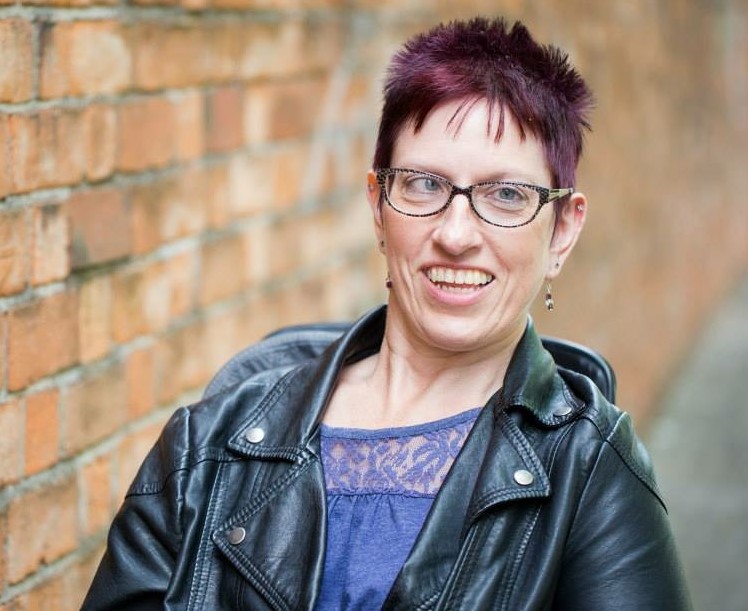 A portrait photo of a woman with short, burgundy hair. She is wearing black glasses, a black leather jacket and a blue top. She is smiling.