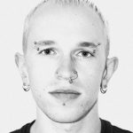 A black and white portrait photo of a man with very short bleached hair. He's wearing a black t-shirt and has several piercings on his face.