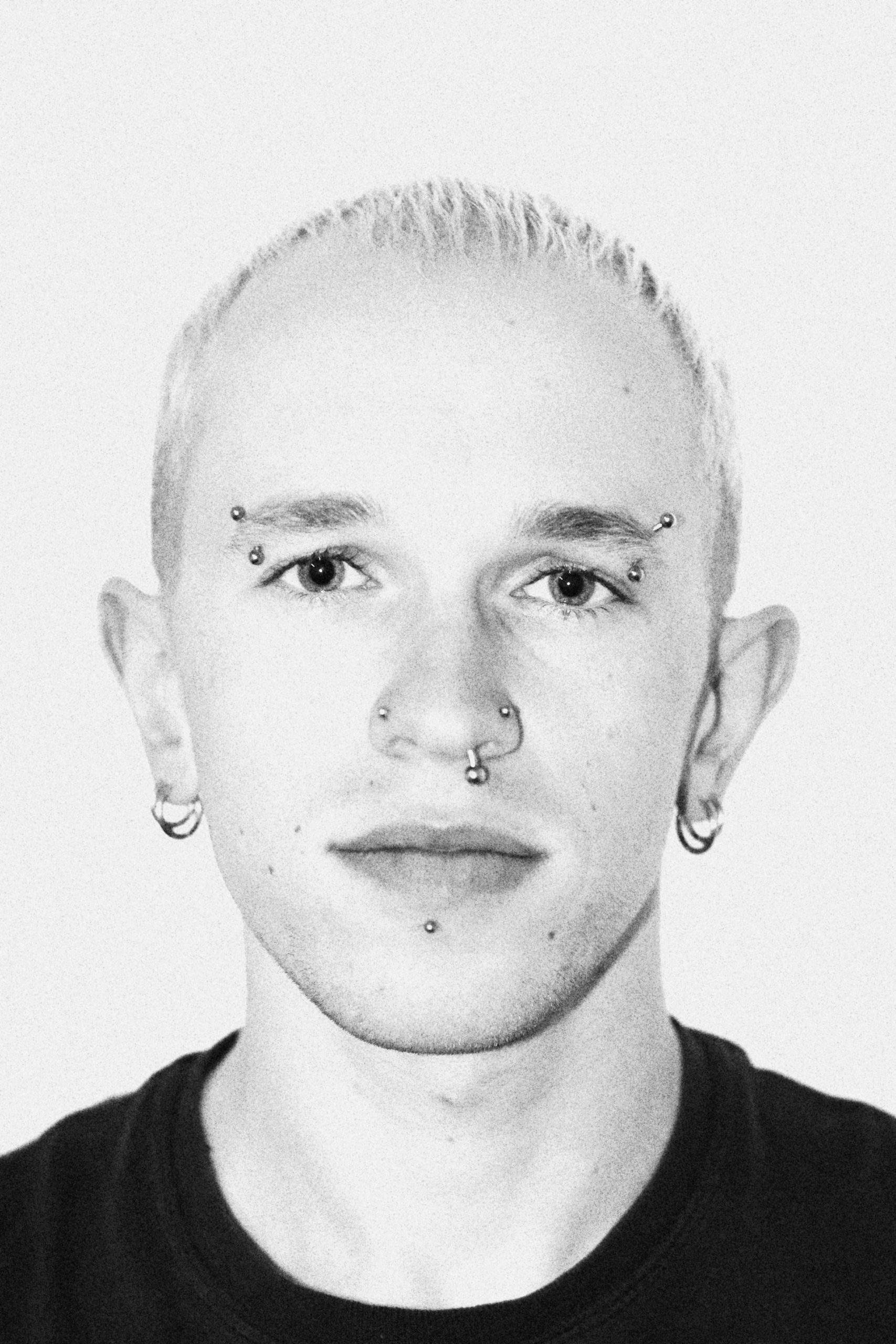 A black and white portrait photo of a man with very short bleached hair. He's wearing a black t-shirt and has several piercings on his face.
