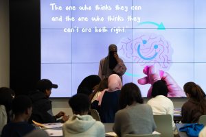 A woman facilitator stands in front of a group of engaged young people. Behind them a screen reads "the one who thinks they can and the one who thinks they can't are both right."