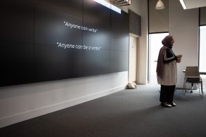 A woman facilitator wearing a lilac hijab and an Adidas top stands in front of a screen reading, "Anyone can write. Anyone can be a writer."