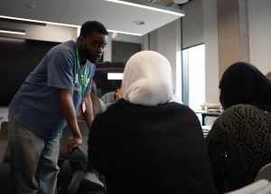 A male facilitator leans over a desk, speaking to two young women.