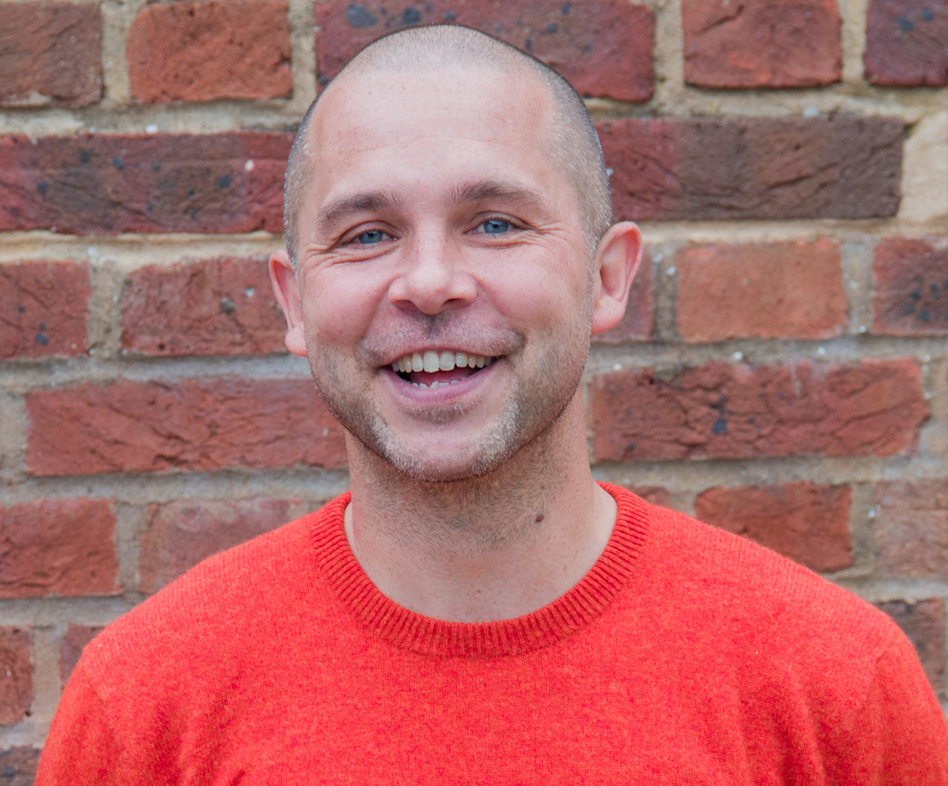 A photographic headshot of a man standing against a brick wall, wearing an orange jumper. He's smiling,