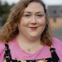 Fat white woman looks at the camera with a half-smile. She has mid-length brown hair in a side parting, and wears a pink top with dungarees with stars and moons on them.