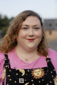 Fat white woman looks at the camera with a half-smile. She has mid-length brown hair in a side parting, and wears a pink top with dungarees with stars and moons on them.