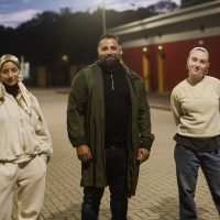 Three actors: Chifaa, Kate and Thaer look at the camera in the car park of the community centre.