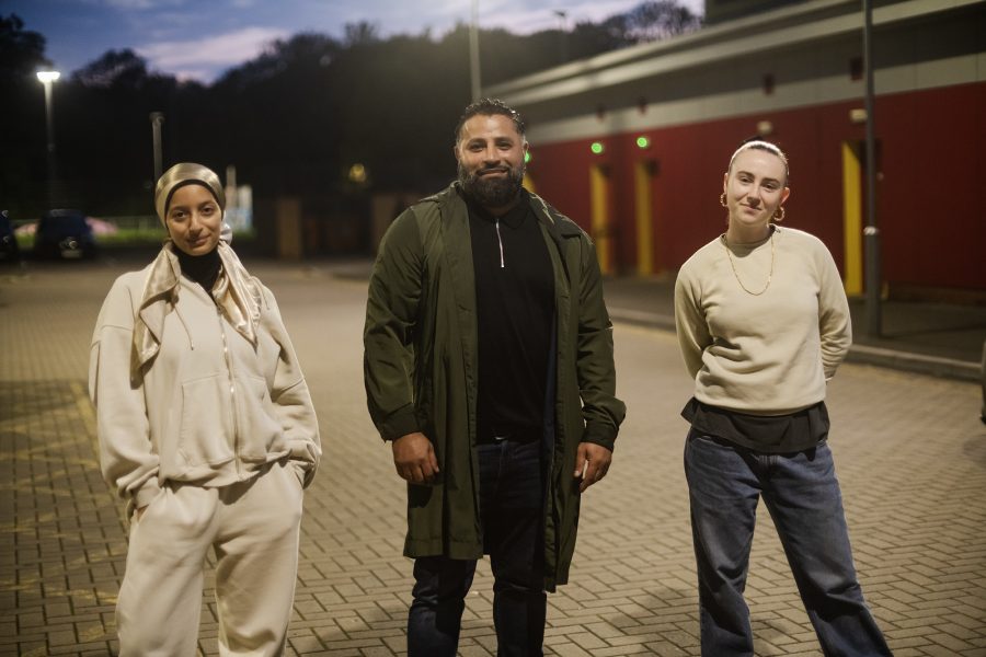 Three actors: Chifaa, Kate and Thaer look at the camera in the car park of the community centre.