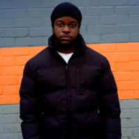 Trustee Fahadi stands against a blue and orange wall. He looks directly into the camera. He is a young Black man wearing a black hat and winter coat.