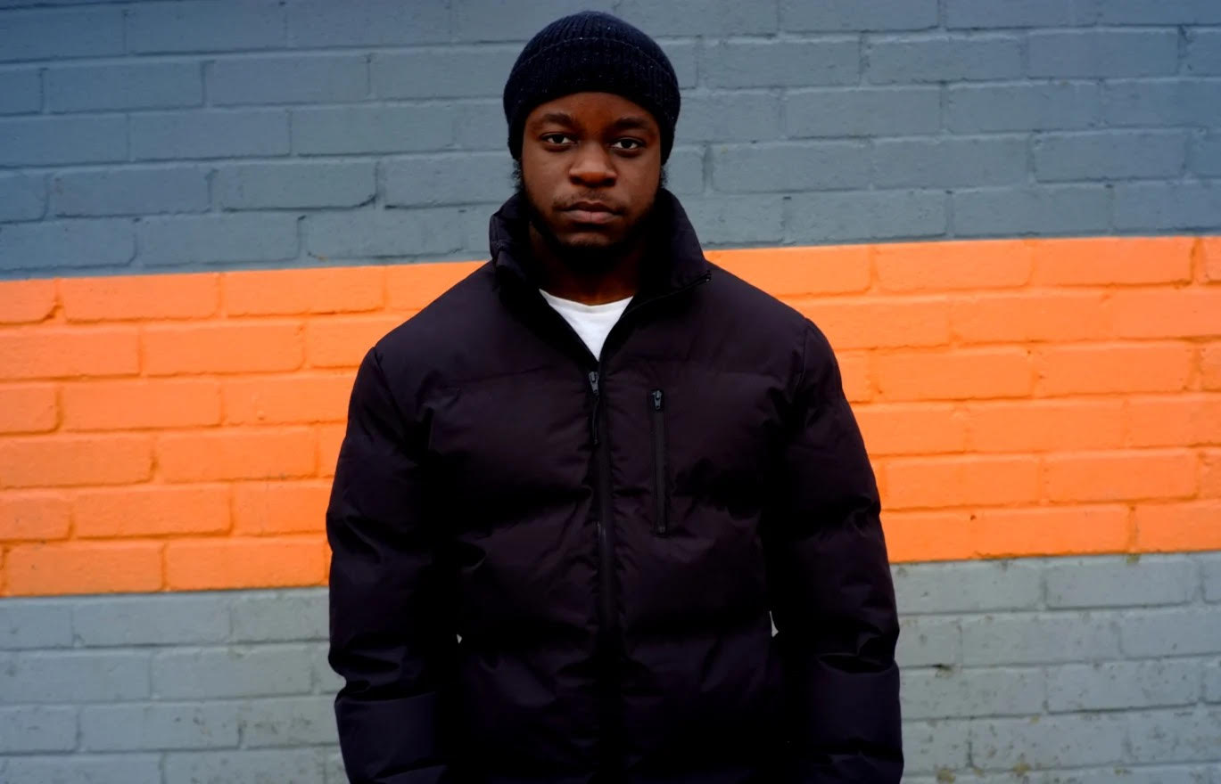 Trustee Fahadi stands against a blue and orange wall. He looks directly into the camera. He is a young Black man wearing a black hat and winter coat.