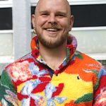 Andrew Westle is a white man with a short light brown beard and no head hair. He is smiling widely and wearing a funky, bright patterned multicoloured fleece.