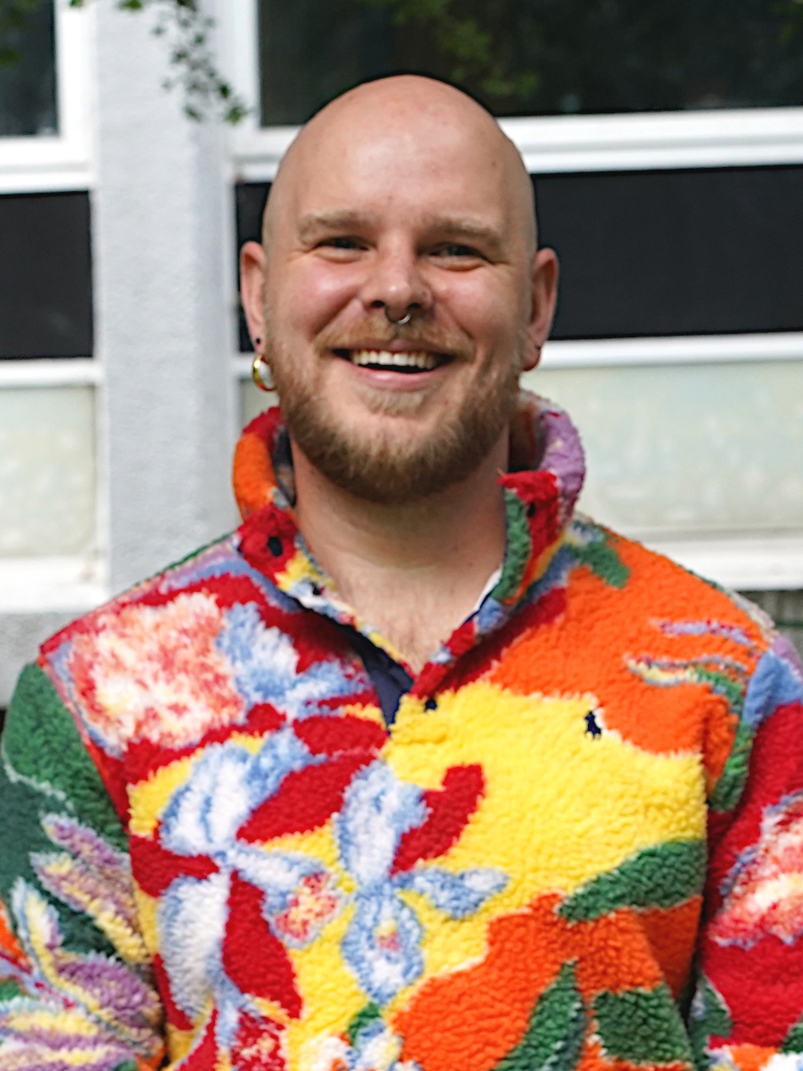 Andrew Westle is a white man with a short light brown beard and no head hair. He is smiling widely and wearing a funky, bright patterned multicoloured fleece.