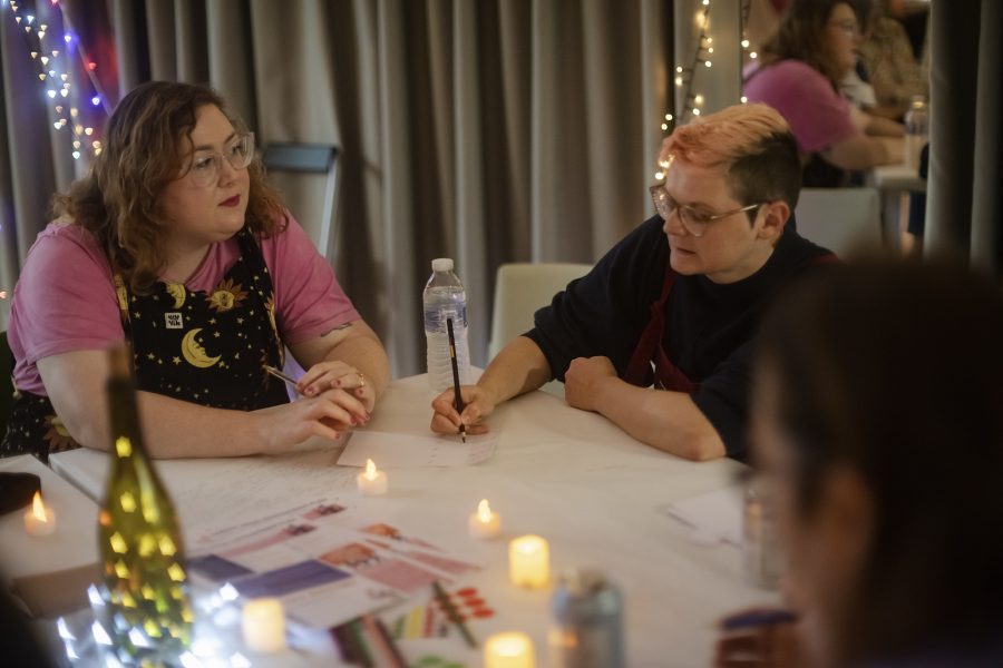 Two women sit at a table with pens and paper. They are surrounded with warm fairy lights and tea lights.