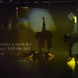 the shadow of a performer is silhouetted against a wall with the words "kill the bill"