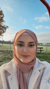 A smiling woman in a baby pink hijab and a white coat, on a sunny cold morning.