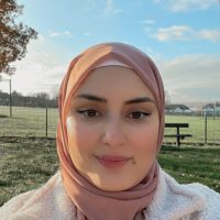 A smiling woman in a baby pink hijab and a white coat, on a sunny cold morning.