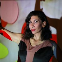Performance Artist Riham, a Palestinian woman with shoulder length dark hair, holds a string of heart shapes. She has red lipstick and is looking enigmatically at the camera.