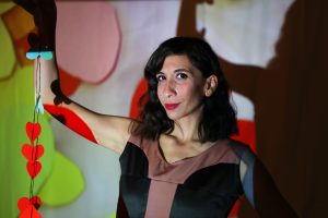 Performance Artist Riham, a Palestinian woman with shoulder length dark hair, holds a string of heart shapes. She has red lipstick and is looking enigmatically at the camera.