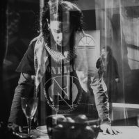 Layered image of the performers. Hussina leans with her weight on her hands, staring at a bowl of apples, while Soul Roberts leans against a door in the background