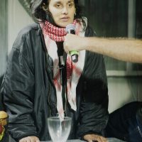 Hussina Raja stands at a table, speaking into a microphone with redacted documents spread on the table beneath her. 